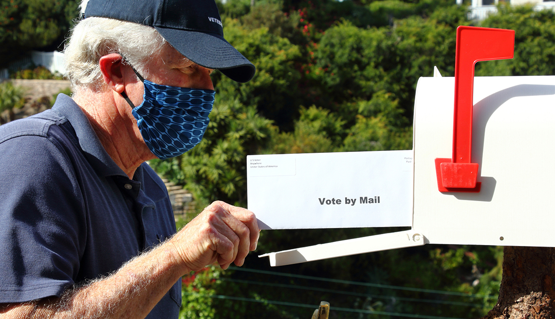 a man wearing a face mask is putting an envelope that says vote by mail on it into a mailbox
